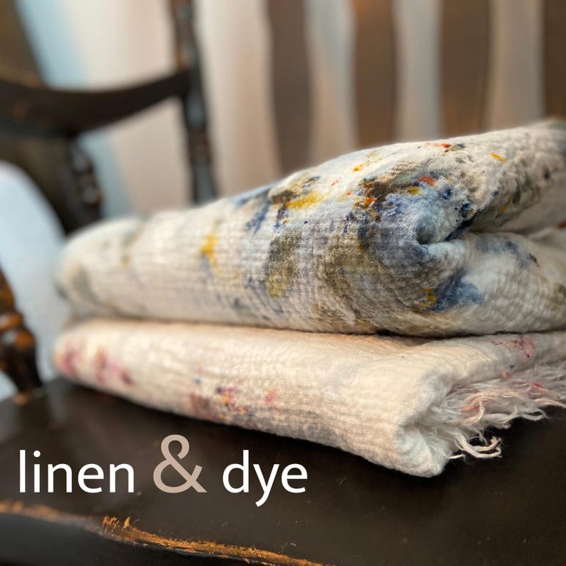 May include: Two folded linen textiles with a textured appearance. The top textile features a pattern of blue, yellow, and green hues, while the bottom one has pink and red accents. The text "linen & dye" is at the bottom.