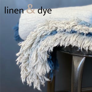 May include: A close-up of a fringed textile with the text "linen & dye". The textile features a combination of white, blue, and gray hues. The fringe hangs over a dark metal surface, creating a textured and layered effect.