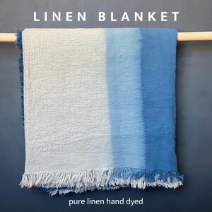 May include: A linen blanket with a two-tone design, transitioning from white to shades of blue. The blanket has a fringed edge and is displayed on a wooden rod. The text "LINEN BLANKET" is at the top, and "pure linen hand dyed" is at the bottom.