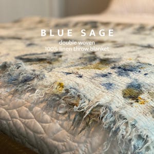 May include: A close-up of a double woven 100% linen throw blanket. The blanket is white with blue and yellow accents. The text "BLUE SAGE" is at the top, with "double woven" and "100% linen throw blanket" below.