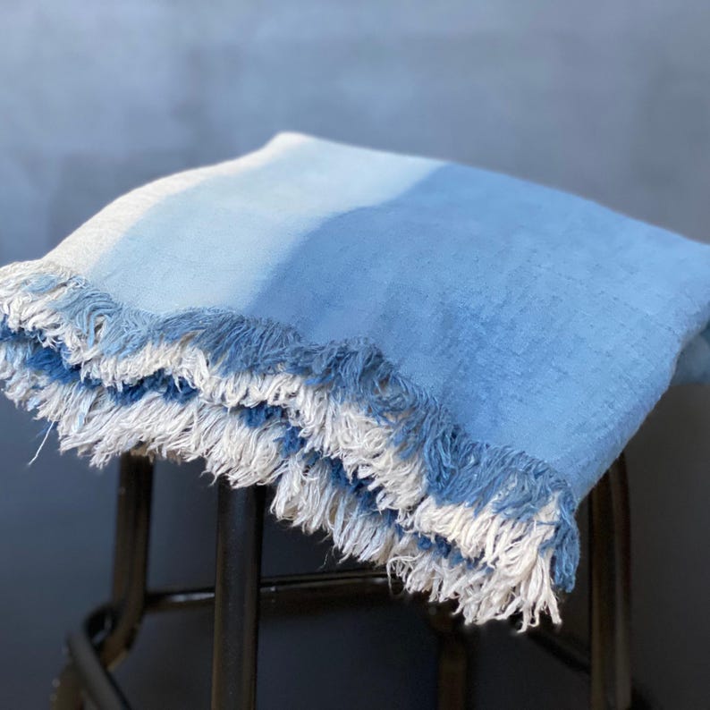 May include: A folded blue throw blanket with white fringe. The blanket has a gradient of light to medium blue hues. The fringe is a mix of white and blue. The blanket is made of a soft, textured material.