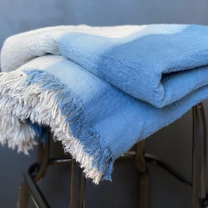 May include: A folded blue and white blanket with fringed edges. The blanket has a gradient of colors, transitioning from white to light blue and then to a darker blue. The blanket is made of a soft, textured material.