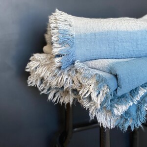 May include: A stack of soft, fringed blankets in shades of blue and gray. The blankets are folded and layered, with the fringed edges visible. The color gradient transitions from light gray to a soft blue. The blankets appear to be made of a woven material.