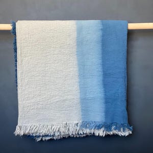 May include: A blue and white ombre throw blanket with fringe. The blanket is hung on a wooden rod against a dark blue wall. The color gradient transitions from white to light blue to a darker blue.
