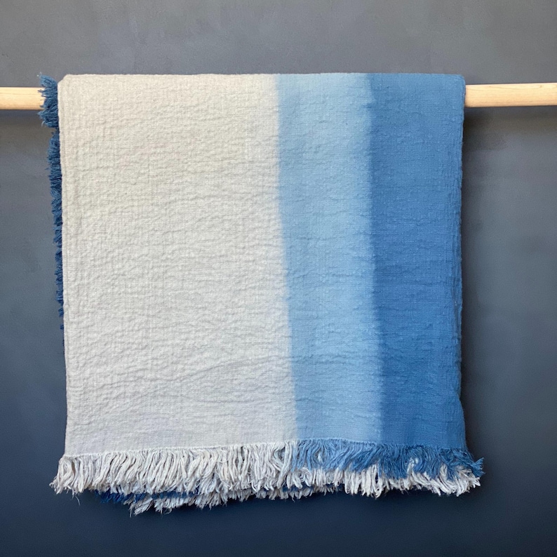 May include: A blue and white ombre throw blanket with fringe. The blanket is hung on a wooden rod against a dark blue wall. The color gradient transitions from white to light blue to a darker blue.
