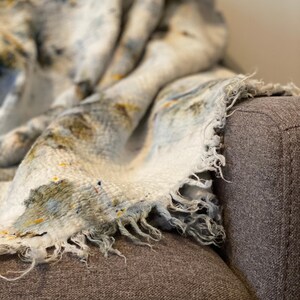 May include: A soft, fringed blanket in shades of white, blue, and yellow, draped over a gray armchair. The blanket has a speckled pattern and a textured appearance, creating a cozy and inviting aesthetic.