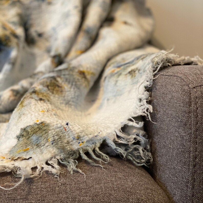 May include: A soft, fringed blanket in shades of white, blue, and yellow, draped over a gray armchair. The blanket has a speckled pattern and a textured appearance, creating a cozy and inviting aesthetic.