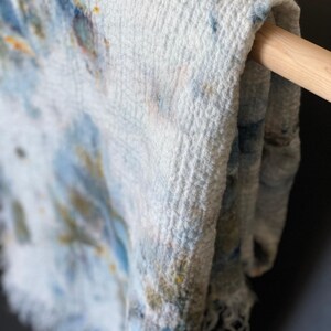 May include: Close-up of a white, textured textile with blue, yellow, and orange watercolor-like patterns. The fabric is draped over a wooden rod, showcasing its soft, fringed edge. The textile appears to be a scarf or a light blanket.