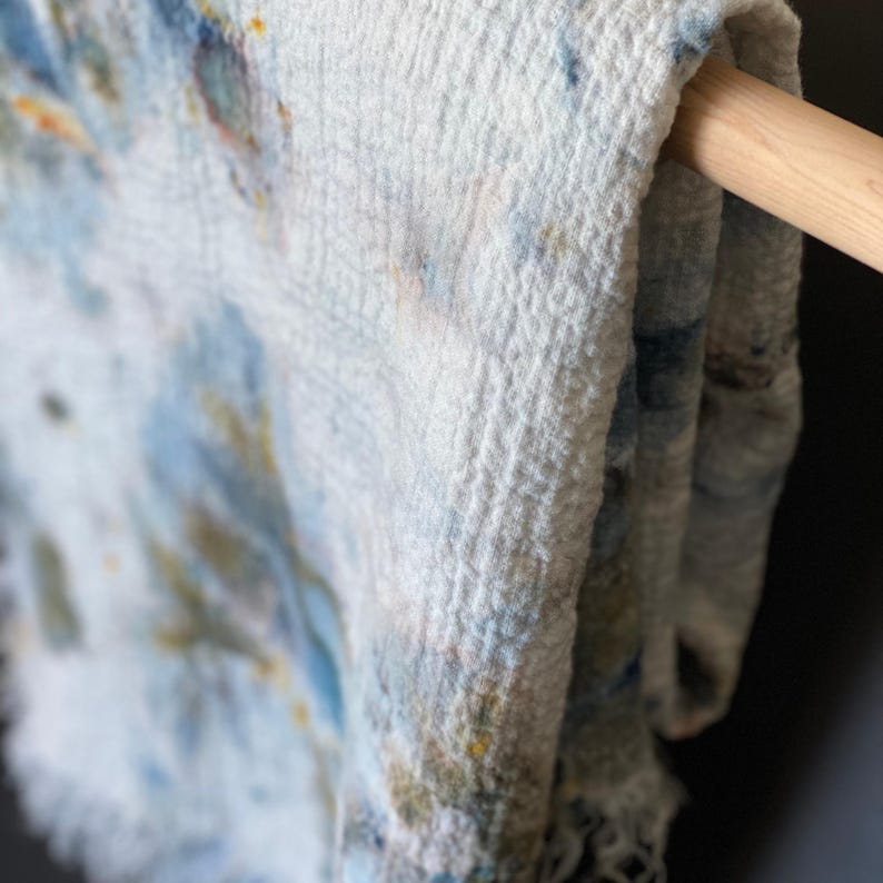 May include: Close-up of a white, textured textile with blue, yellow, and orange watercolor-like patterns. The fabric is draped over a wooden rod, showcasing its soft, fringed edge. The textile appears to be a scarf or a light blanket.