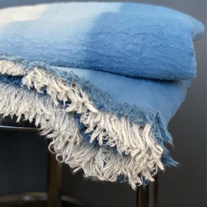May include: A folded blue blanket with a gradient effect, transitioning from a darker shade to a lighter hue. The blanket features a fringe of white and blue threads along the edge, adding texture and detail to the soft textile.