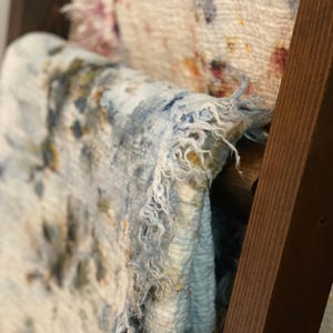 May include: Close-up of a light blue textile with a frayed edge, featuring dark blue and yellow splotches. The fabric is draped over a wooden frame, with another textile visible in the background.