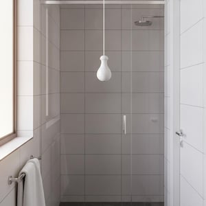 May include: A white pendant light with a bulbous shape hangs in a bathroom featuring white tiled walls, a glass shower enclosure with a silver handle, a dark grey tiled floor, and a white towel hanging on a silver rack near a window.