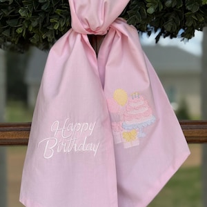 May include: A light pink fabric with the embroidered text "Happy Birthday" in white script. The fabric features an embroidered cake design with yellow, pink, and blue accents. The fabric is tied in a knot.