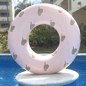 May include: A light pink inflatable pool float with a repeating pattern of hand illustrations giving the middle finger. The float is circular and is resting on a white stone surface above a blue swimming pool.