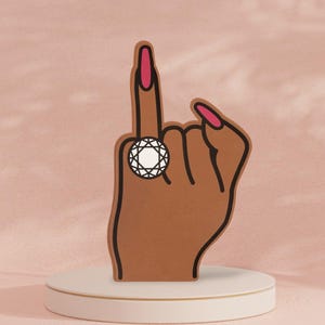 May include: A brown hand with pink fingernails is making a 'middle finger' gesture. The hand is wearing a ring with a large, clear diamond.