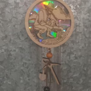 May include: A wooden ornament with a holographic image of a person panning for gold. The ornament is hanging from a brown cord with a wooden bead and two wooden tools. There are three black beads at the bottom of the cord.