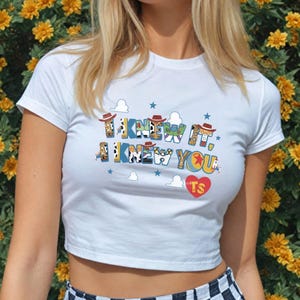May include: A white short-sleeved crop top featuring the text I KNEW IT, I KNEW YOU in colorful patterned letters with small cowboy hats on top, blue stars, white clouds, and a red heart with the letters TS printed on the front of the shirt.