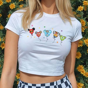 May include: White crop top featuring cartoon character balloon designs. The balloons are in various colors and feature iconic character faces. The shirt has a crew neck and short sleeves. The background is a wall of yellow flowers.