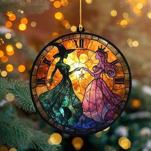 May include: Round Christmas ornament with a stained-glass design. The ornament features a witch and a princess holding hands, set against a warm, golden background. The design is framed by a clock face with Roman numerals. A gold string is attached for hanging.
