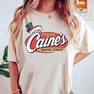 May include: A cream-colored short-sleeve t-shirt featuring a graphic of a cartoon character wearing a top hat with teeth visible. The text reads Amazing Caine's DIGITAL CIRCUS in white, yellow, and black letters over a red oval background.