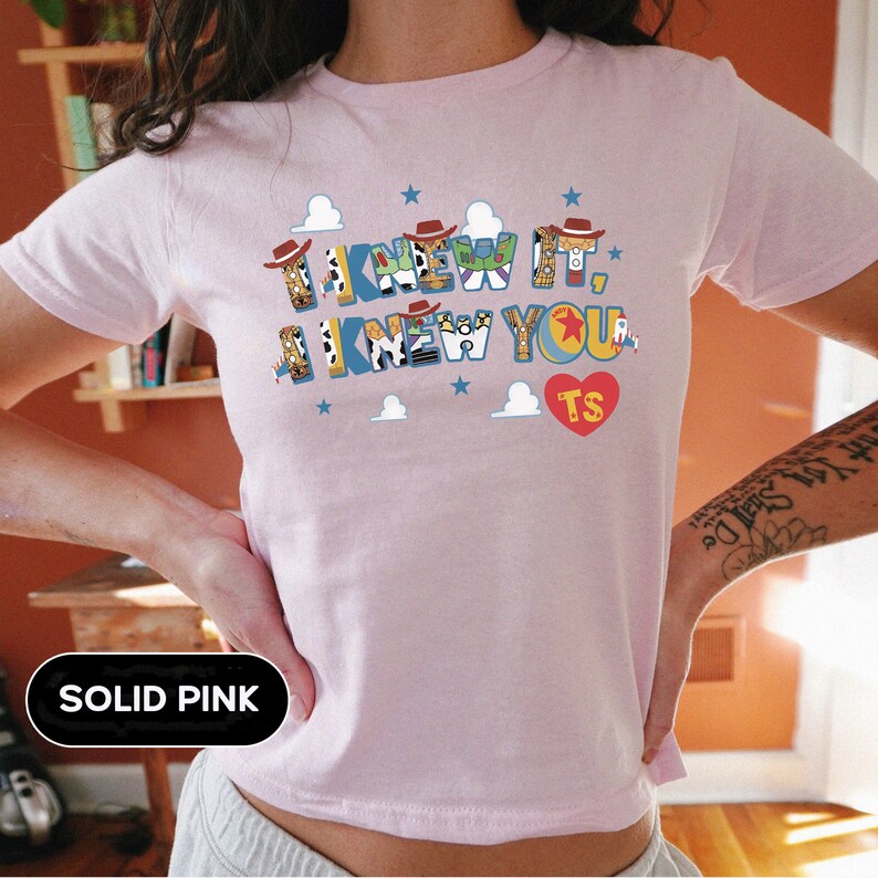 May include: A light pink short-sleeved t-shirt featuring a graphic print with the text I KNEW IT, I KNEW YOU. The letters are decorated with patterns inspired by cowboy and space ranger characters, small brown hats, blue stars, white clouds, and a red heart with the letters TS.