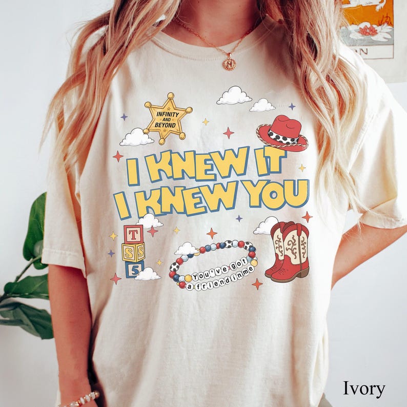 May include: An ivory t-shirt featuring a graphic print with the text "I KNEW IT I KNEW YOU". The design includes a yellow sheriff star badge with "INFINITY AND BEYOND", a red cowboy hat, alphabet blocks, a beaded bracelet with "YOU'VE GOT A FRIEND IN ME", and red cowboy boots.