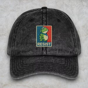 May include: A distressed black baseball cap featuring an embroidered patch with a green frog wearing a blue scarf. The patch is set against a red and blue background, with the word "RESIST" in white.