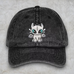 May include: A black denim baseball cap with a white embroidered cartoon dragon with blue eyes. The dragon is sitting on its hind legs with its wings spread out.