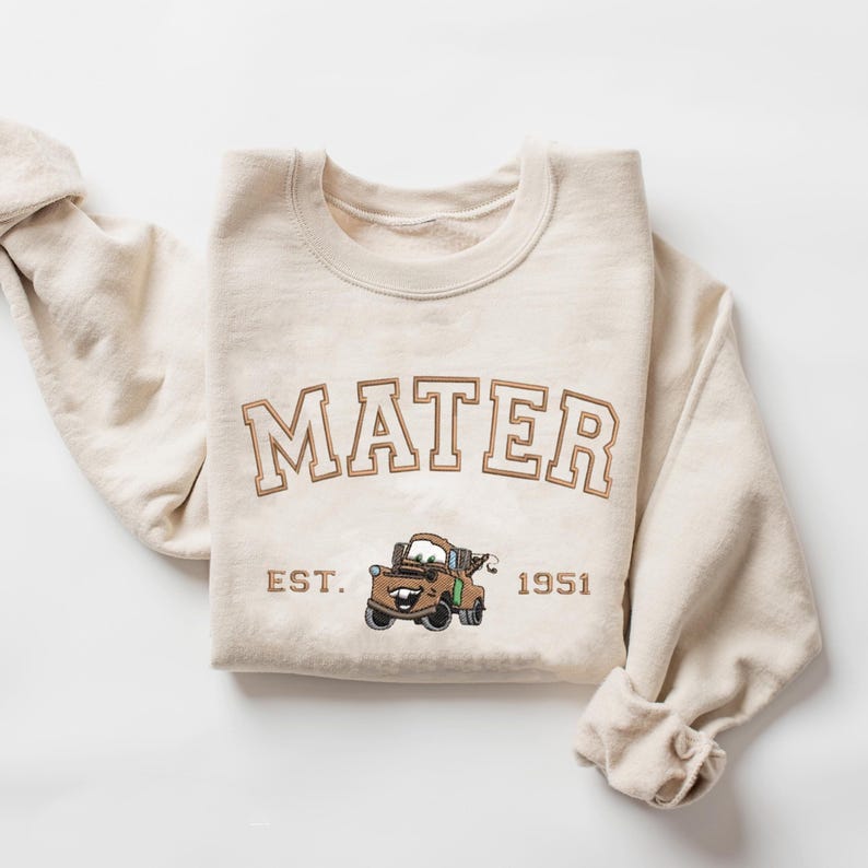Puede incluir: Una sudadera beige con la palabra "MATER" en letras marrones con un contorno blanco. Debajo de la palabra hay una imagen de dibujos animados de Mater de la pel&iacute;cula Cars y el texto "EST. 1951".