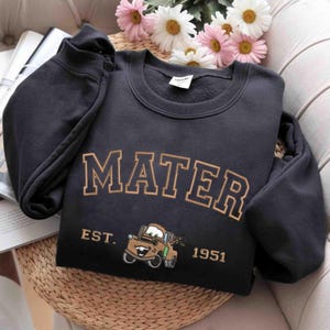 Puede incluir: Sudadera negra con el texto "Mater Est. 1951" en letras doradas. Debajo del texto hay una imagen de dibujos animados de Mater de la pel&iacute;cula Cars.