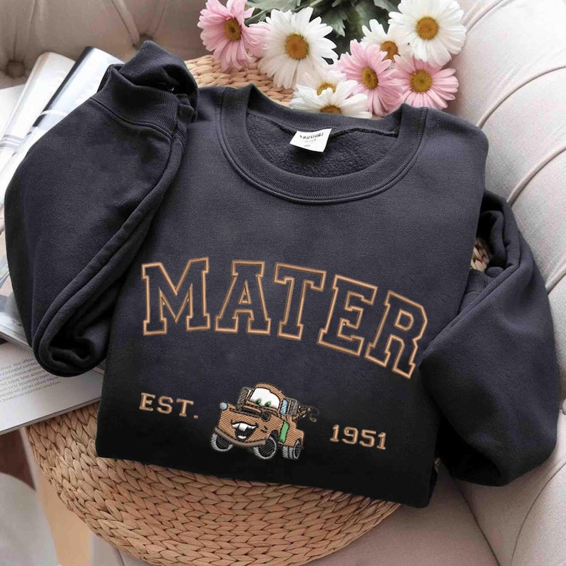 Puede incluir: Sudadera negra con el texto "Mater Est. 1951" en letras doradas. Debajo del texto hay una imagen de dibujos animados de Mater de la pel&iacute;cula Cars.