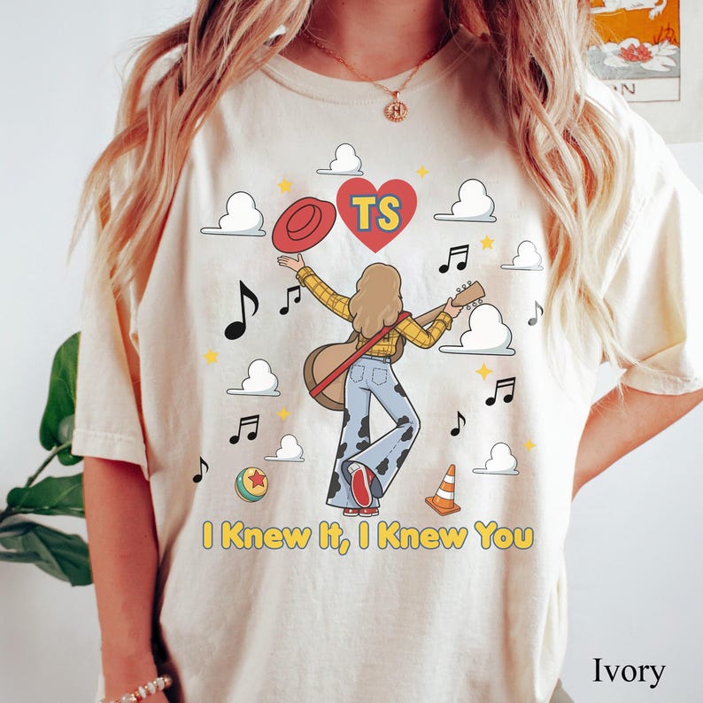 May include: An ivory t-shirt featuring a graphic of a person with long hair playing a guitar, wearing a yellow plaid shirt and cow-print trousers. The design includes a red hat, a red heart with the letters TS, clouds, music notes, a ball, and a traffic cone, with the text I Knew It, I Knew You printed below.