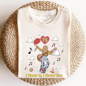 May include: A cream-coloured t-shirt featuring a graphic of a person with long hair playing a guitar, wearing a yellow plaid shirt and cow-print trousers. The design includes a red hat, a heart with the letters TS, musical notes, clouds, a ball, and a cone, with the text "I Knew It, I Knew You" at the bottom.