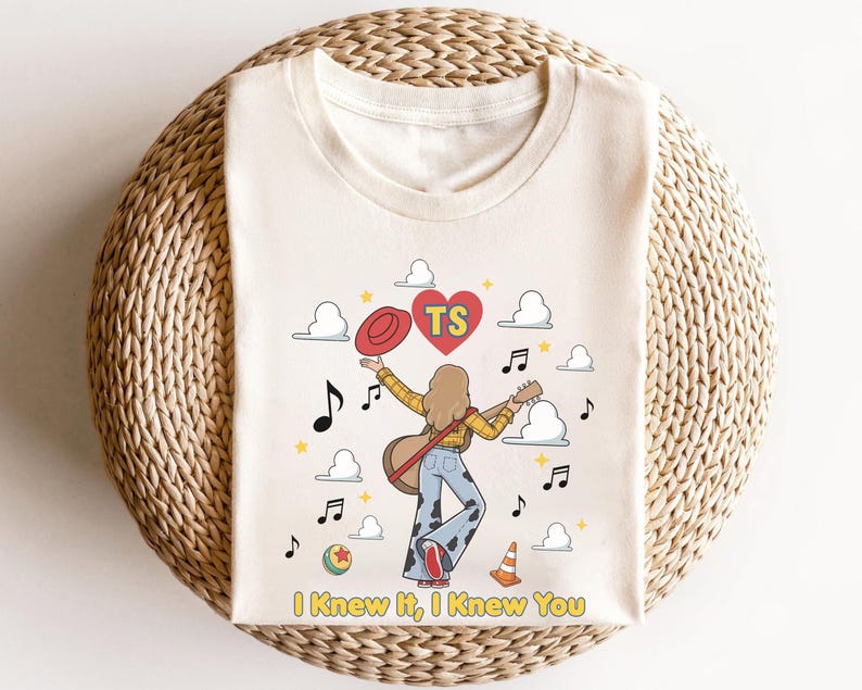 May include: A cream-coloured t-shirt featuring a graphic of a person with long hair playing a guitar, wearing a yellow plaid shirt and cow-print trousers. The design includes a red hat, a heart with the letters TS, musical notes, clouds, a ball, and a cone, with the text "I Knew It, I Knew You" at the bottom.