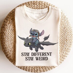 Pu&ograve; includere: T-shirt color crema con un'illustrazione a cartoni animati di Stitch che cavalca un drago. Stitch indossa gli occhiali. Il testo "STAY DIFFERENT STAY WEIRD" &egrave; stampato in nero sotto l'immagine.