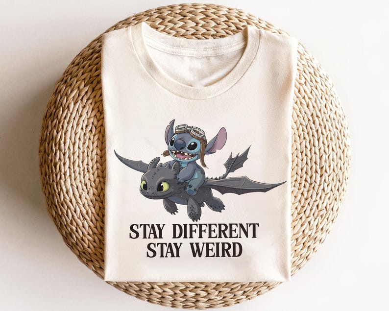 Pu&ograve; includere: T-shirt color crema con un'illustrazione a cartoni animati di Stitch che cavalca un drago. Stitch indossa gli occhiali. Il testo "STAY DIFFERENT STAY WEIRD" &egrave; stampato in nero sotto l'immagine.