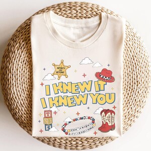 May include: A cream-coloured t-shirt featuring the text I KNEW IT I KNEW YOU in yellow with blue outlines. Graphics include a yellow sheriff star badge, a red cowboy hat, a stack of alphabet blocks, a beaded bracelet, and red cowboy boots.