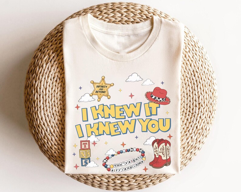May include: A cream-coloured t-shirt featuring the text I KNEW IT I KNEW YOU in yellow with blue outlines. Graphics include a yellow sheriff star badge, a red cowboy hat, a stack of alphabet blocks, a beaded bracelet, and red cowboy boots.