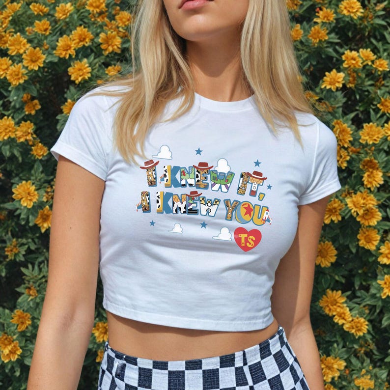 May include: A white short-sleeved crop top featuring the text I KNEW IT, I KNEW YOU in colorful patterned letters with small cowboy hats on top, blue stars, white clouds, and a red heart with the letters TS printed on the front of the shirt.