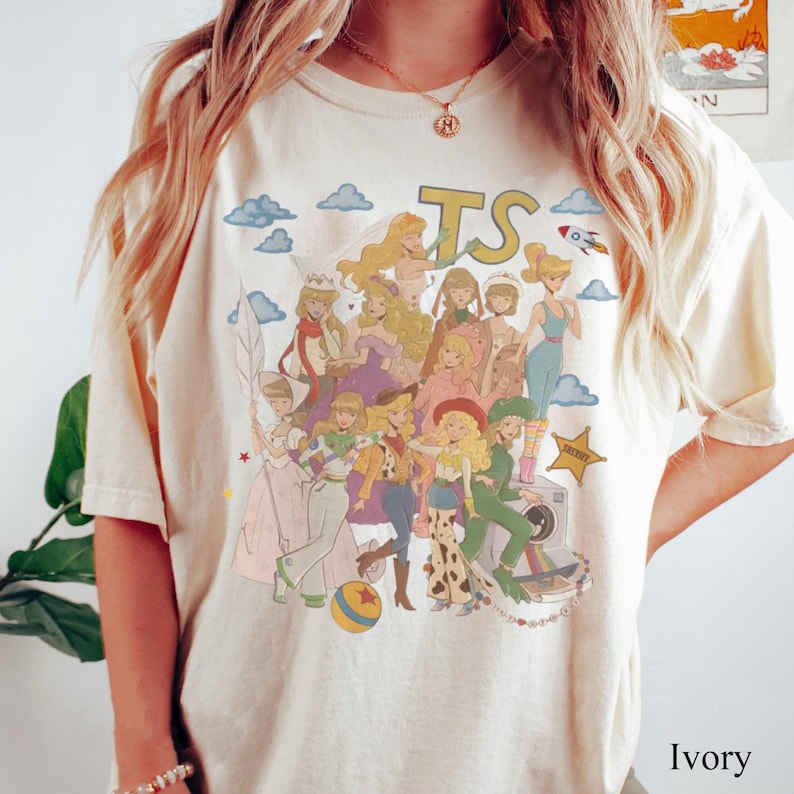 Może przedstawiać: T-shirt w kolorze kości słoniowej z nadrukiem graficznym przedstawiającym wiele animowanych postaci w stylu kolażu. Projekt zawiera litery TS, rakietę, ż&oacute;łtą gwiazdę, piłkę i aparat fotograficzny. Postacie są przedstawione w r&oacute;żnych strojach.
