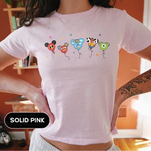 May include: Solid pink t-shirt featuring a whimsical design of cartoon character balloons. The balloons are in various colors and patterns, with star accents. The shirt is a light pink color.