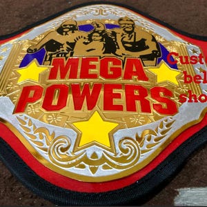 Könnte beinhalten: Ein goldener und roter Wrestling-Meisterschaftsgürtel mit den Worten "MEGA POWERS" in Rot. Der Gürtel hat eine silberne und goldene Mittelplatte mit gelben Sternen und einen schwarzen Riemen. Der Text "Custom belt shop" ist ebenfalls sichtbar.
