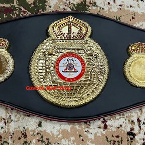May include: A gold boxing championship belt with a black leather strap and a large central plate featuring a globe, two boxers, and the World Boxing Association logo. The belt has two smaller plates with crowns on either side of the central plate.