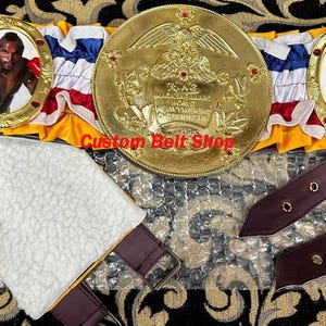 May include: A gold boxing championship belt with a red, white and blue ribbon. The belt has two round medallions with images of boxers and a large central medallion with an eagle and the text "World Heavyweight Champion".