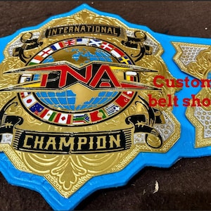 May include: A custom championship belt with a blue, gold, and black design. The belt features the text "INTERNATIONAL CHAMPION" and "TNA" with a globe graphic and various flags. The text "Custom belt shop" is also visible.