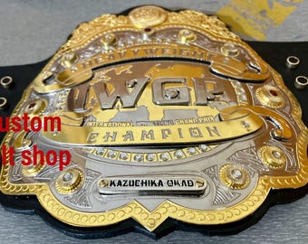May include: A gold and silver championship belt with the text "IWGP Heavyweight Champion" and "International Wrestling Grand Prix" on it. The belt also has the name "KAZUCHIKA OKADA" on it.