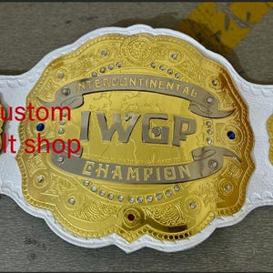 Könnte beinhalten: Ein weißer Wrestling-Championschaftsgürtel mit goldenen Akzenten. Der Gürtel trägt den Schriftzug "Intercontinental IWGP Champion".