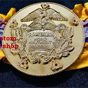May include: A gold championship belt, inscribed with "Rocky Balboa World Heavyweight Champion." The belt has an eagle design and decorative features with red accents. It is set against a yellow and blue backdrop.