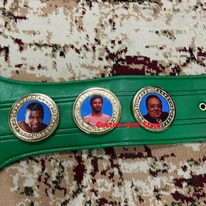 May include: A green boxing championship belt with gold accents and three circular medallions featuring portraits of boxers. The medallions have the words "World Boxing Council" inscribed around the edges.