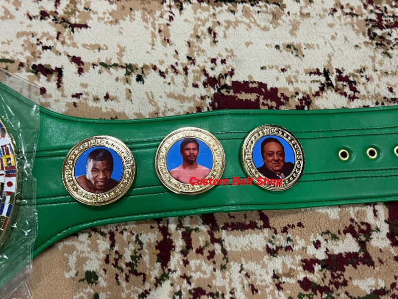 May include: A green boxing championship belt with gold accents and three circular medallions featuring portraits of boxers. The medallions have the words "World Boxing Council" inscribed around the edges.